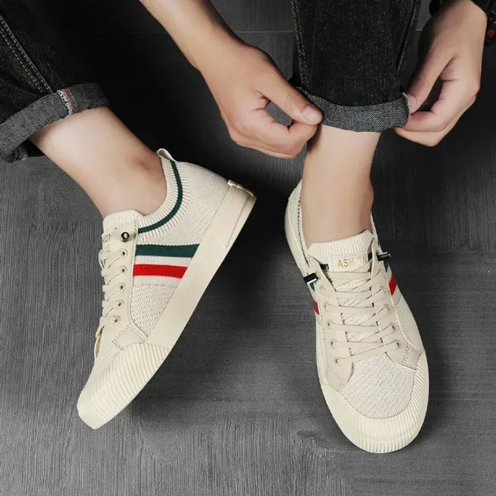 A person wearing beige sneakers with a breathable mesh upper and red and green stripes is rolling up the cuff of their dark jeans while sitting on a dark floor.