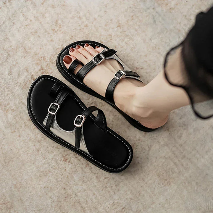 A person wearing black open-toe sandals with a leather upper and dual buckle design stands on a light, textured surface. One sandal is on the foot, while the other rests nearby.