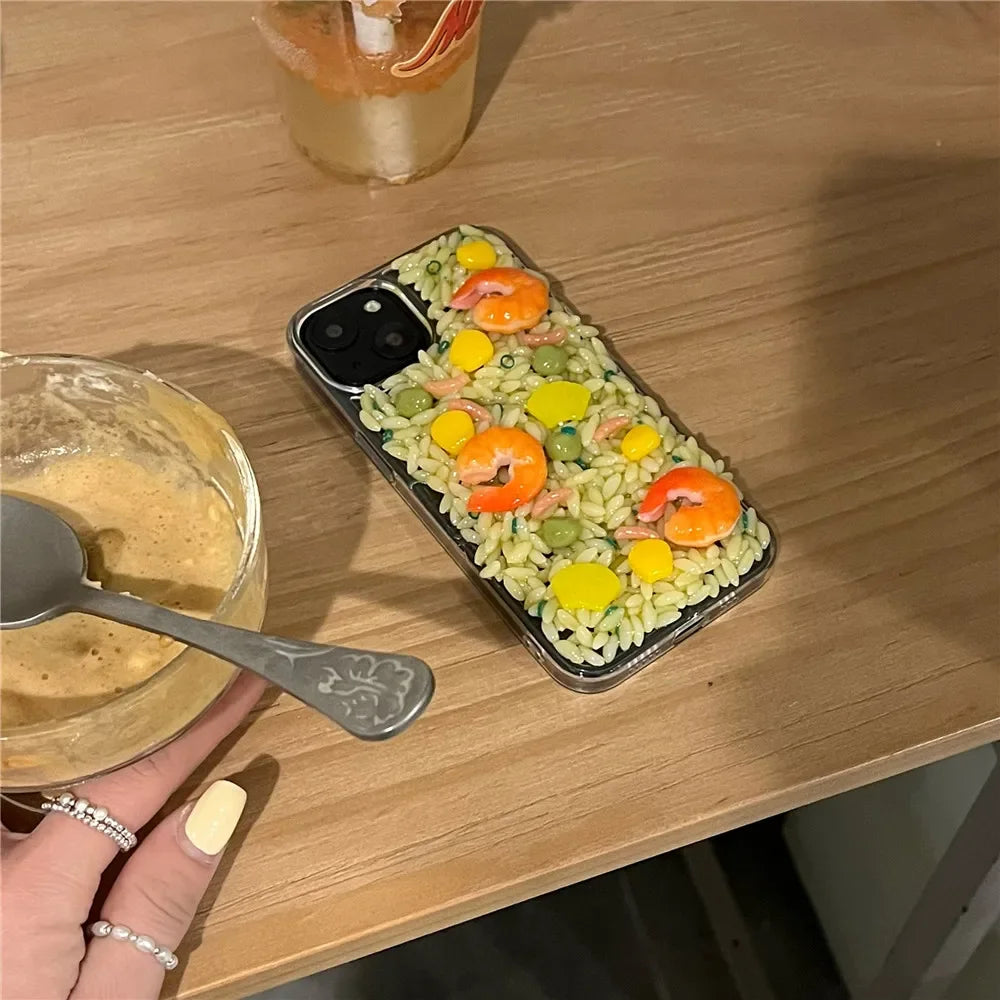 A phone with rugged protection features sits on a wooden table, its case decorated with 3D detailing to resemble a rice dish with shrimp, peas, and corn. Nearby are a glass of drink and a bowl held by a hand with painted nails.
