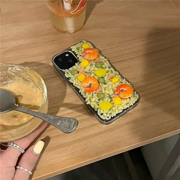 A phone with rugged protection features sits on a wooden table, its case decorated with 3D detailing to resemble a rice dish with shrimp, peas, and corn. Nearby are a glass of drink and a bowl held by a hand with painted nails.