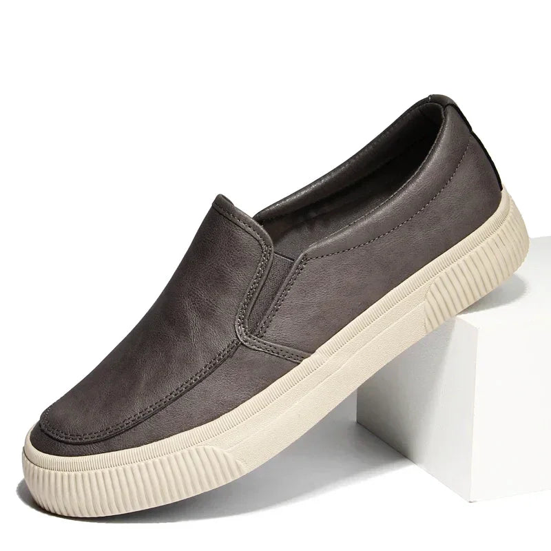 A single gray men’s slip-on sneaker with a hybrid rubber sole and white textured finish is displayed, propped at an angle on a white block against a plain background.
