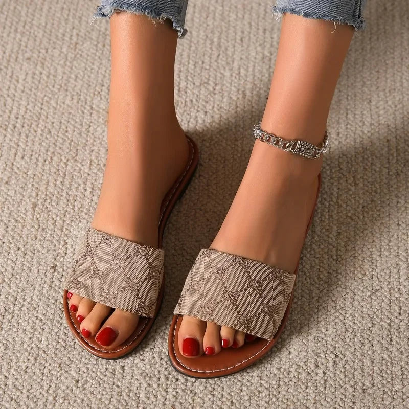 A person wearing timeless shoes in a slide silhouette and a silver anklet stands on beige carpet. Their toenails are painted red, and they are wearing light blue frayed hem jeans.