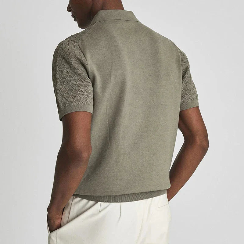 A person wearing a short-sleeved, light olive green wrinkle-resistant herringbone polo with textured sleeves and cream pants, shown from the back with hands in their pockets.