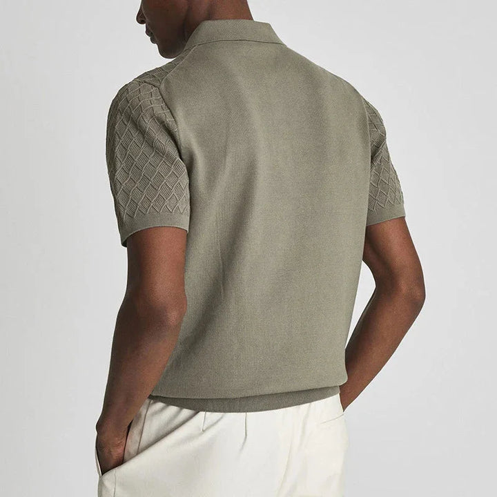 A person wearing a short-sleeved, light olive green wrinkle-resistant herringbone polo with textured sleeves and cream pants, shown from the back with hands in their pockets.