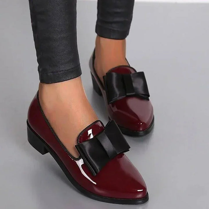 A person wearing shiny burgundy patent leather loafers with a large statement bow on the front, paired with black skinny pants and standing on a gray surface.