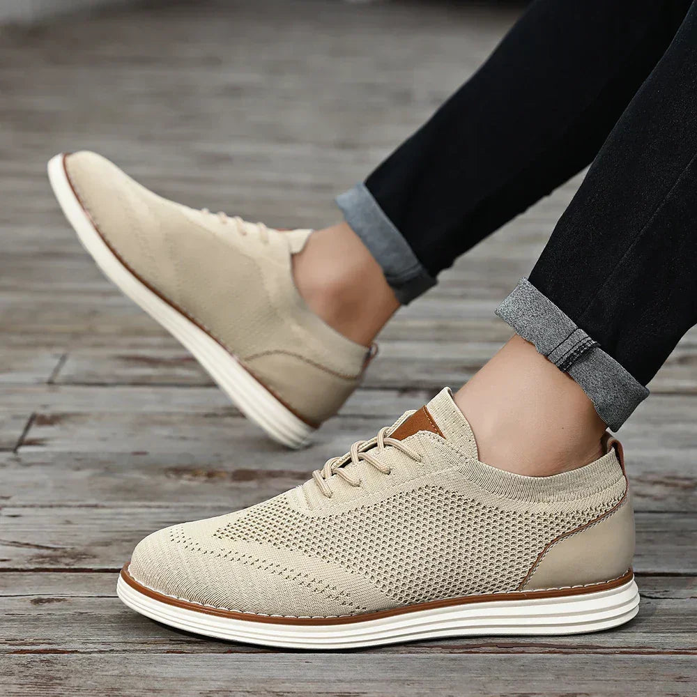 A person wearing breathable knit sneakers with white soles and tan accents stands on a wooden floor. Their jeans, cuffed slightly above the ankles, add to the effortless casual style.