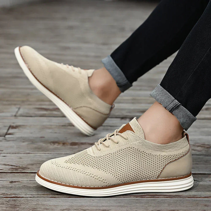 A person wearing breathable knit sneakers with white soles and tan accents stands on a wooden floor. Their jeans, cuffed slightly above the ankles, add to the effortless casual style.