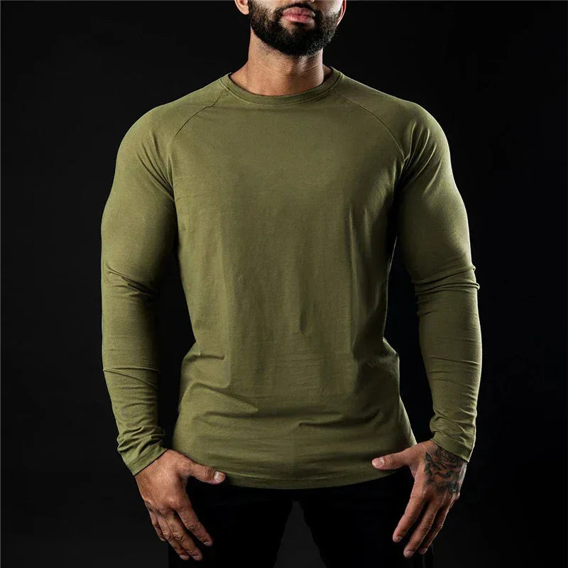 A man in a fitted, long sleeve stretch workout shirt stands against a black background with his hands on his hips. His face is partially out of the frame.