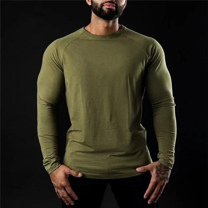 A man in a fitted, long sleeve stretch workout shirt stands against a black background with his hands on his hips. His face is partially out of the frame.