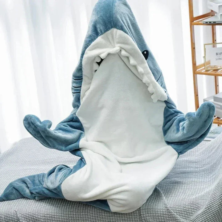 A plush blanket designed to look like a blue and white shark with an open mouth, lying on a bed with light bedding in a bright room. The sharks head and fins give it the playful look of shark pajamas, creating a cozy, inviting vibe.