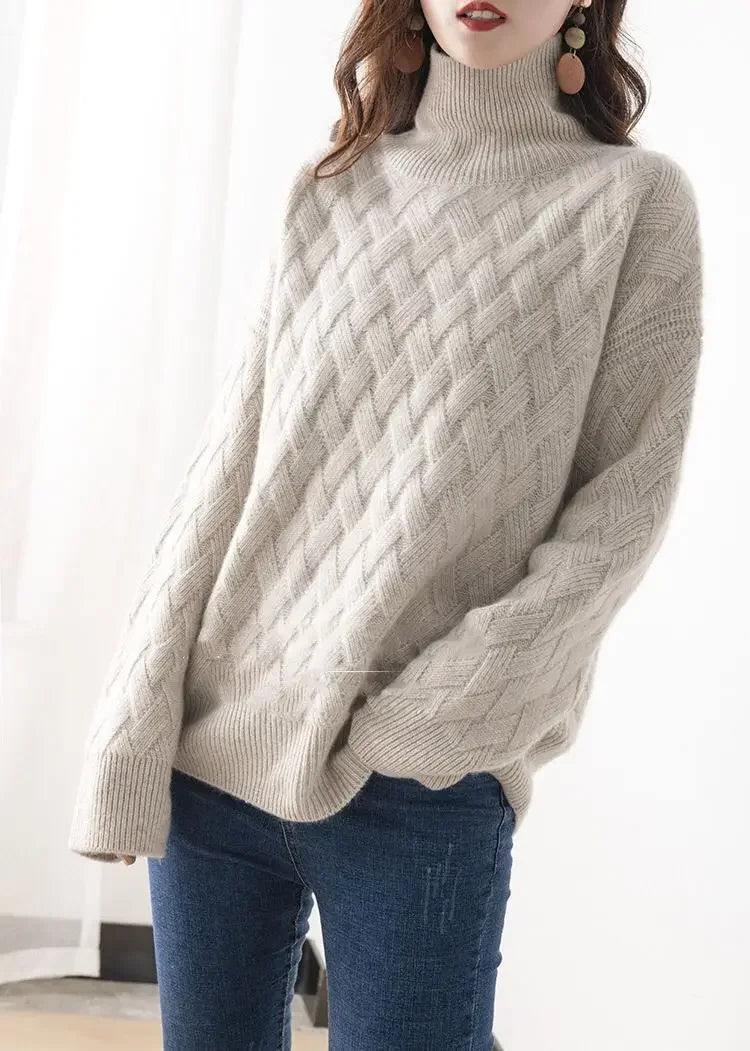 A woman wearing an ultra soft, light beige cable-knit turtleneck sweater and blue jeans stands indoors, hands relaxed at her sides. She has long wavy hair and is wearing large round earrings.