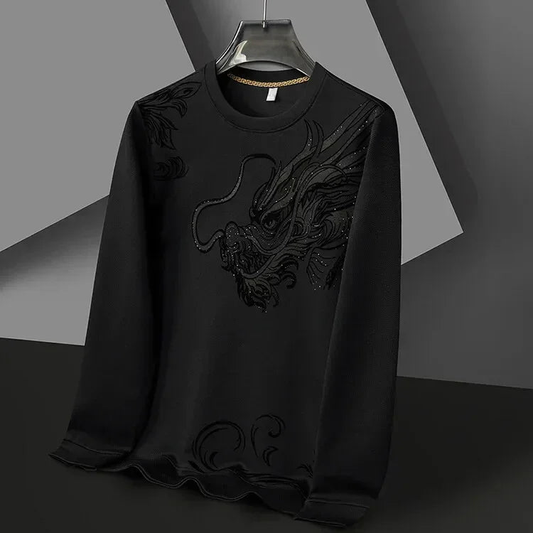 A black long-sleeve Year of the Dragon crewneck hangs on a hanger. It features a textured, embroidered dragon design on the front, blending subtly with ultra-soft cotton fabric. The background is composed of geometric shapes in grey and black.