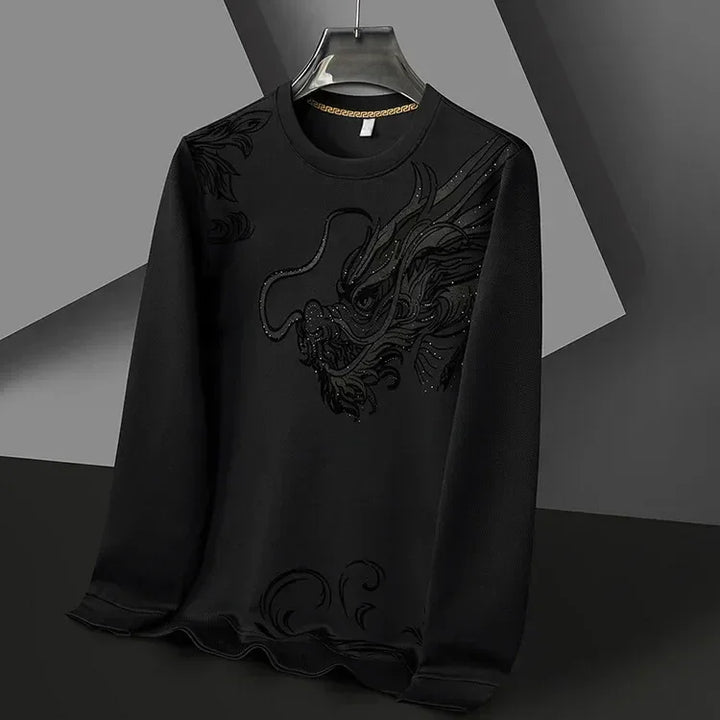A black long-sleeve Year of the Dragon crewneck hangs on a hanger. It features a textured, embroidered dragon design on the front, blending subtly with ultra-soft cotton fabric. The background is composed of geometric shapes in grey and black.