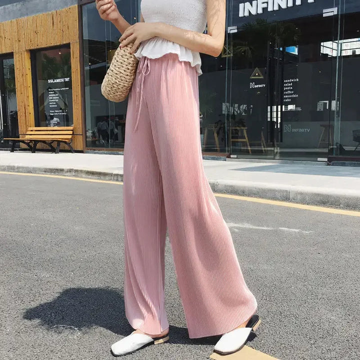 A person wearing pleated ice silk wide leg pants, a white sleeveless top, white shoes, and carrying a woven bag stands on a city street near modern buildings.