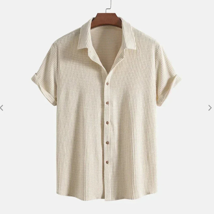 A short-sleeve, cream-colored Bayside Shirt featuring a textured button-up design and collar, crafted from a breathable cotton blend, displayed on a wooden hanger against a plain background.