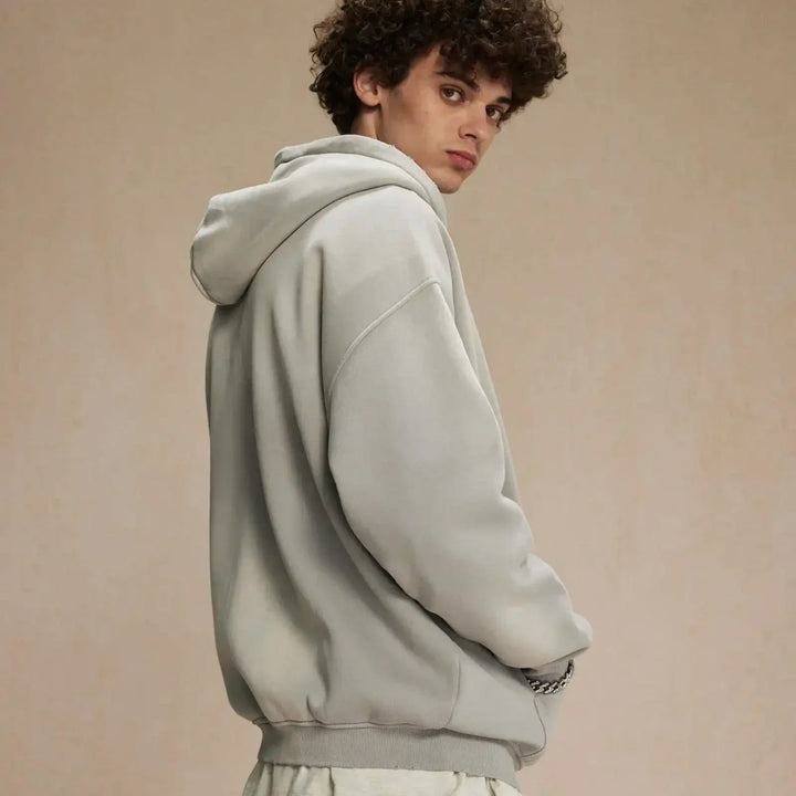 A person with curly hair wearing a relaxed fit, light gray hoodie stands sideways, looking over their shoulder against a neutral beige background.