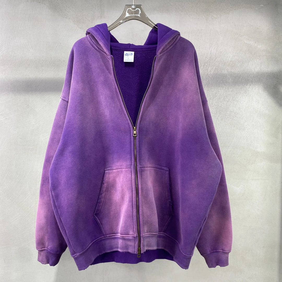 A purple zip-up hoodie with a spray-dyed vintage, faded effect and front pockets, made from thickened fabric, hangs on a clear hanger against a light gray textured wall.