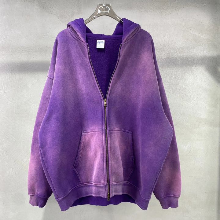 A purple zip-up hoodie with a spray-dyed vintage, faded effect and front pockets, made from thickened fabric, hangs on a clear hanger against a light gray textured wall.