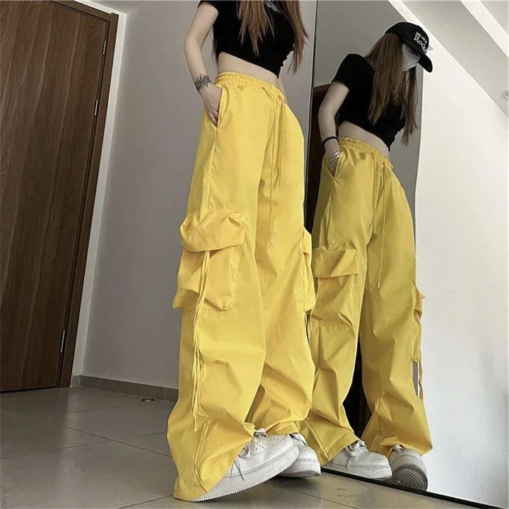 A person wearing loose yellow cargo pants with cargo pockets and a black crop top, white sneakers, and a black cap stands in front of a mirror, showing their reflection in a brightly lit room with a wooden door.