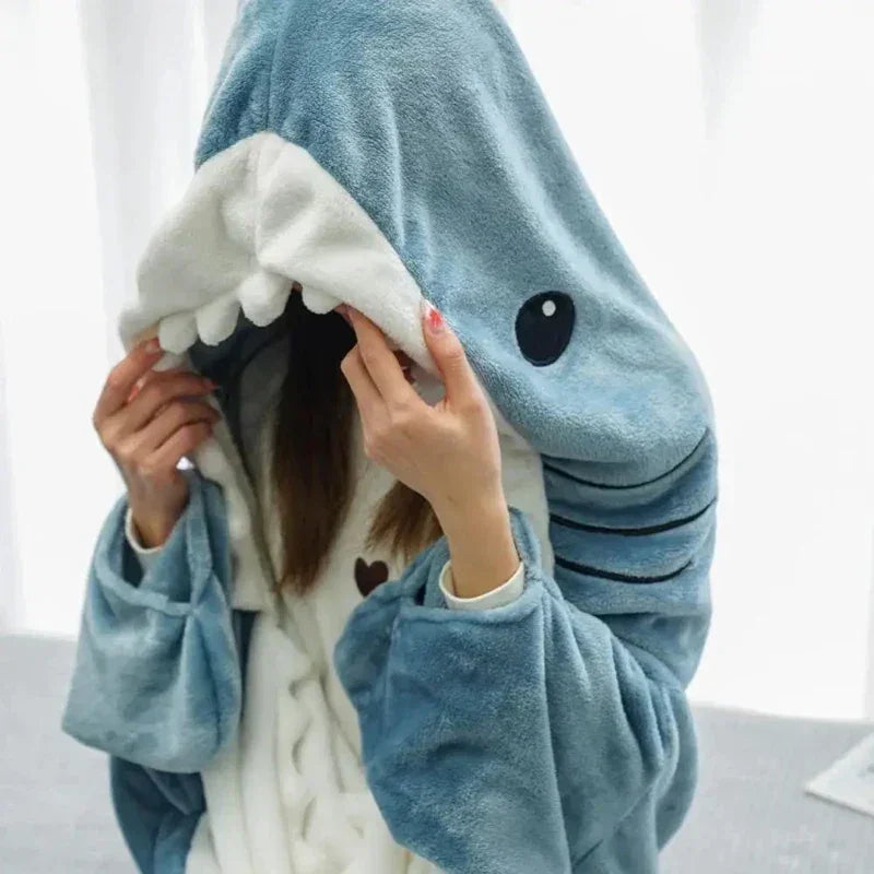 A person wearing cozy shark pajamas with a hood shaped like a shark’s head, covering most of their face, while holding the hood with both hands. The costume features fins, gills, and cartoon-like eyes.