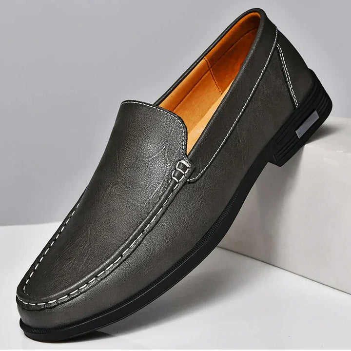 A single dark grey genuine leather loafer with visible white stitching, a rounded toe, and a black rubber sole. This classic slip-on features a light tan inner lining and is displayed on a white surface.
