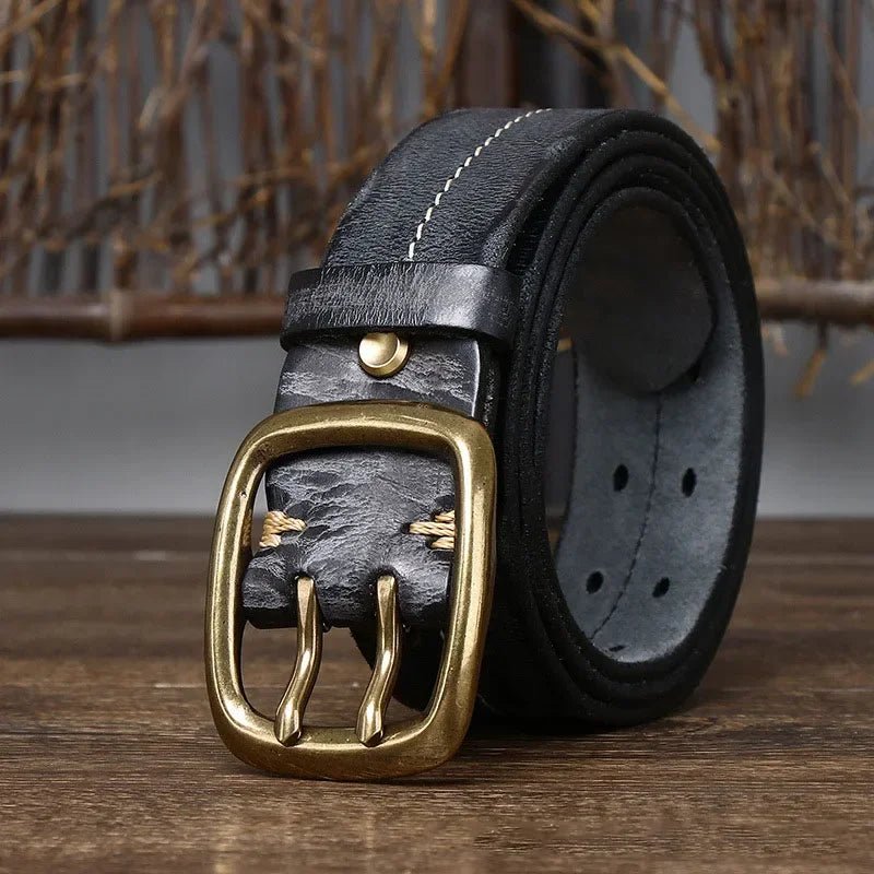 A rolled-up genuine leather belt with visible stitching and a gold-colored double-prong buckle, displayed on a wooden surface with a rustic background, showcasing heritage craftsmanship.