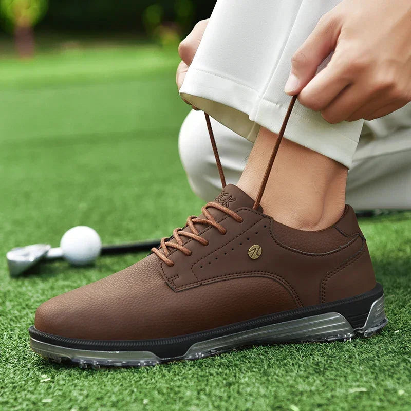 A person ties the brown laces of a brown golf shoe with a high-traction outsole while standing on green grass, with a golf club and ball visible in the background.