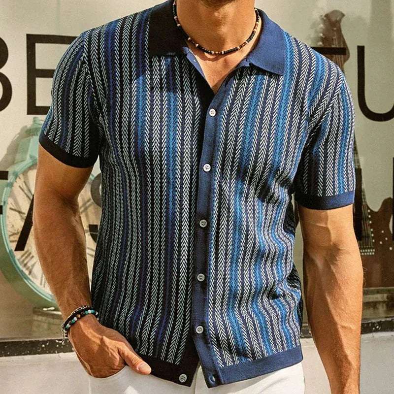 A man wearing a short-sleeve, button-up shirt in a blue and white jacquard herringbone pattern stands indoors. He accessorizes with a beaded necklace and bracelet, with a large clock visible in the background.