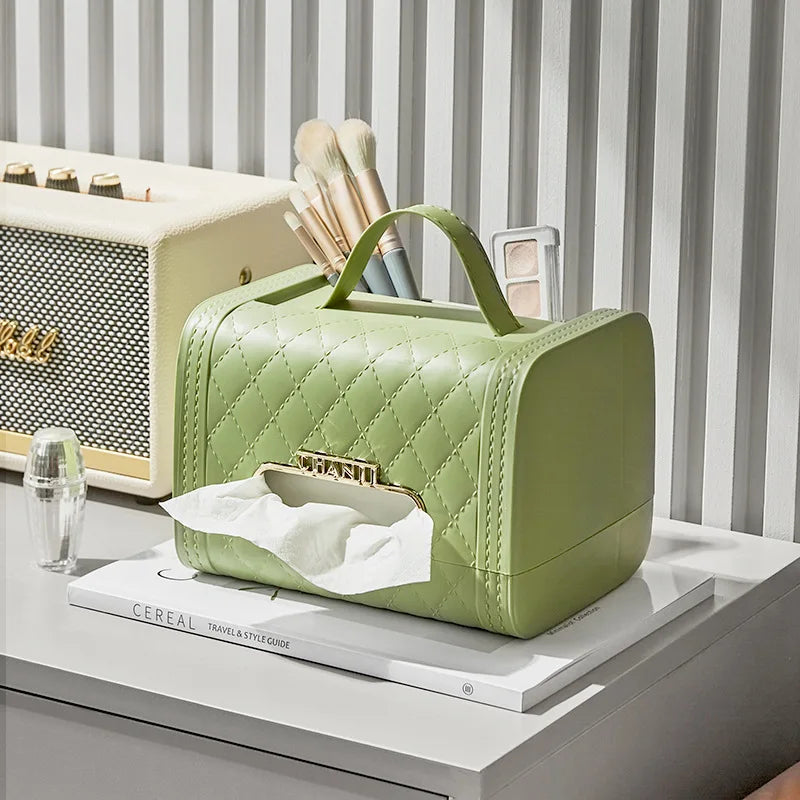 A light green quilted tissue box holder with a handle sits on a white tabletop alongside makeup brushes, a speaker, and a magazine. This stylish home decor piece doubles as an elegant storage solution for tissues. A tissue is partially pulled out in front.