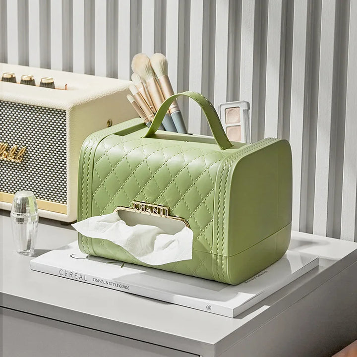 A light green quilted tissue box holder with a handle sits on a white tabletop alongside makeup brushes, a speaker, and a magazine. This stylish home decor piece doubles as an elegant storage solution for tissues. A tissue is partially pulled out in front.