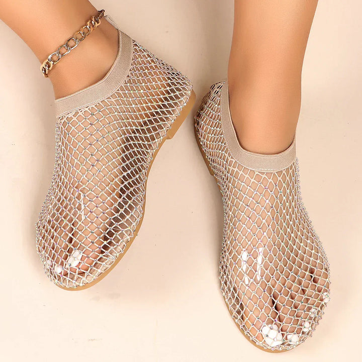 A pair of feet wearing mesh overlay shoes with beige trim and a chain anklet on one ankle, displayed on a light-colored surface.