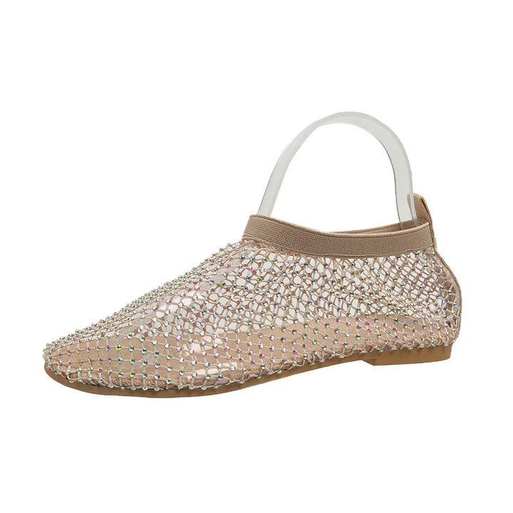 A tan flat shoe with a clear mesh upper and dazzling rhinestone accents, featuring a closed toe, cushioned footbed, tan sole, and an elastic back strap for extra support.