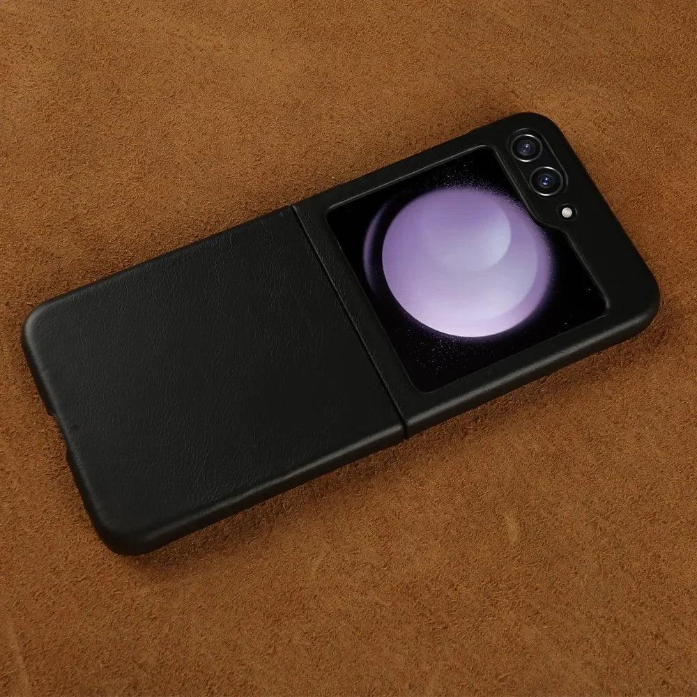 A black Samsung Galaxy Z Flip foldable smartphone with dual rear cameras is partially open, resting on a brown textured surface. The external display shows a circular, glowing graphic. Features shockproof protection for added durability.