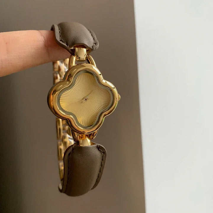 A hand holds a luxury gold-plated clover dial watch, featuring a wavy textured face with no numerals. The timepiece is paired with a genuine leather strap and a gold chain-link band.
