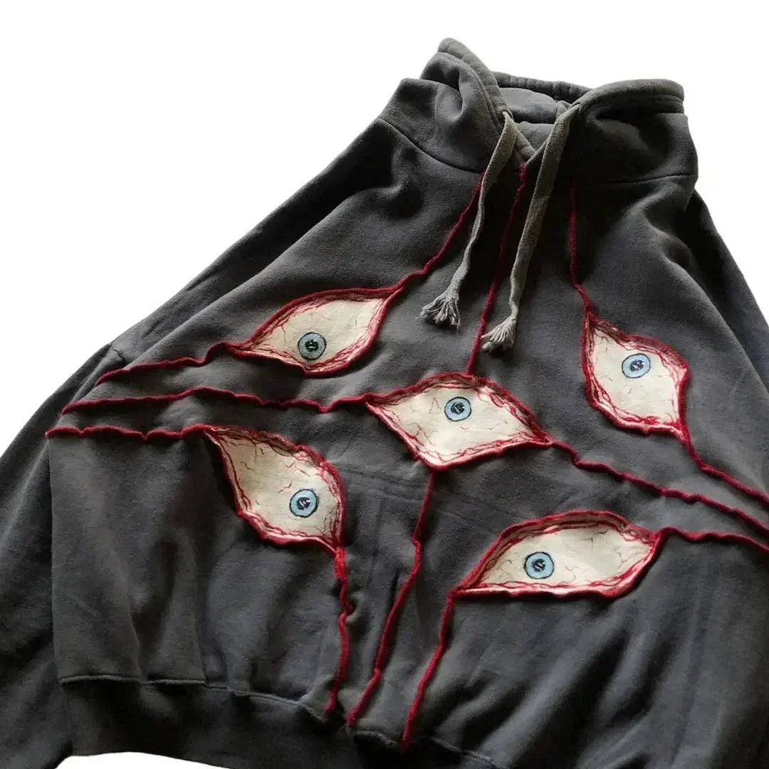A dark gray hoodie featuring an eye pattern patchwork—five blue-irised patches outlined in red stitching across the front. Made from breathable fabric with a comfortable loose fit, it also has light gray drawstrings.