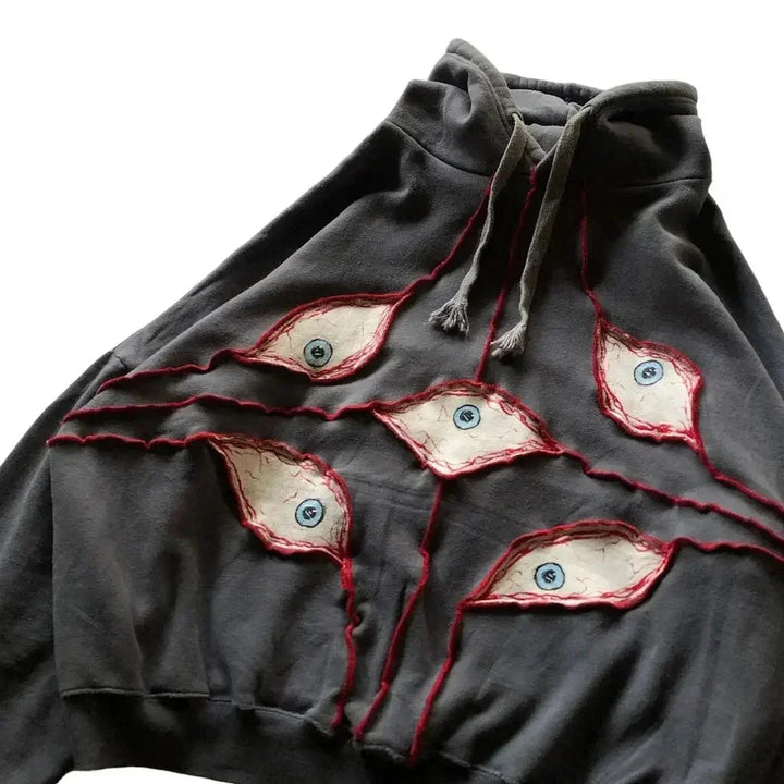 A dark gray hoodie featuring an eye pattern patchwork—five blue-irised patches outlined in red stitching across the front. Made from breathable fabric with a comfortable loose fit, it also has light gray drawstrings.