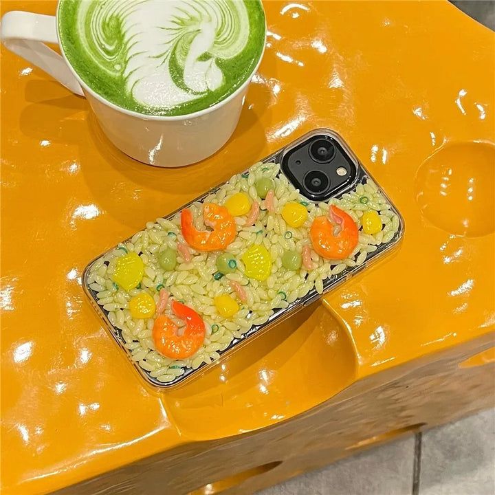 A phone with rugged shock-absorbent protection and 3D detailing that resembles shrimp fried rice sits next to a cup of green matcha latte with latte art, both placed on a bright yellow table.