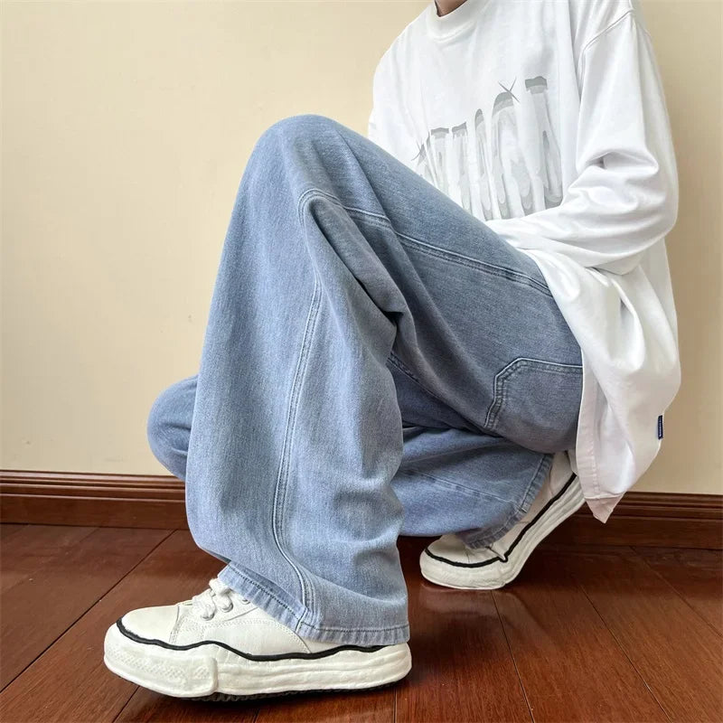 A person wearing relaxed fit, light blue jeans, a white graphic t-shirt, and white sneakers is squatting on a wooden floor against a beige wall. The persons face is not visible.