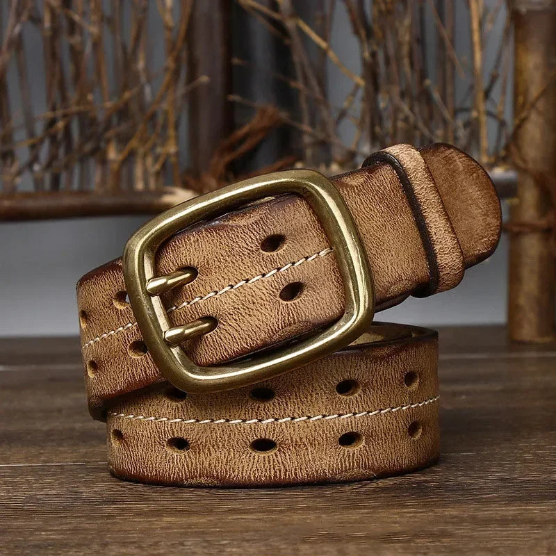A wide, light brown genuine leather belt with double rows of oval holes and white stitching, featuring heritage craftsmanship and a large brass buckle, is coiled on a wooden surface with rustic branches in the background.