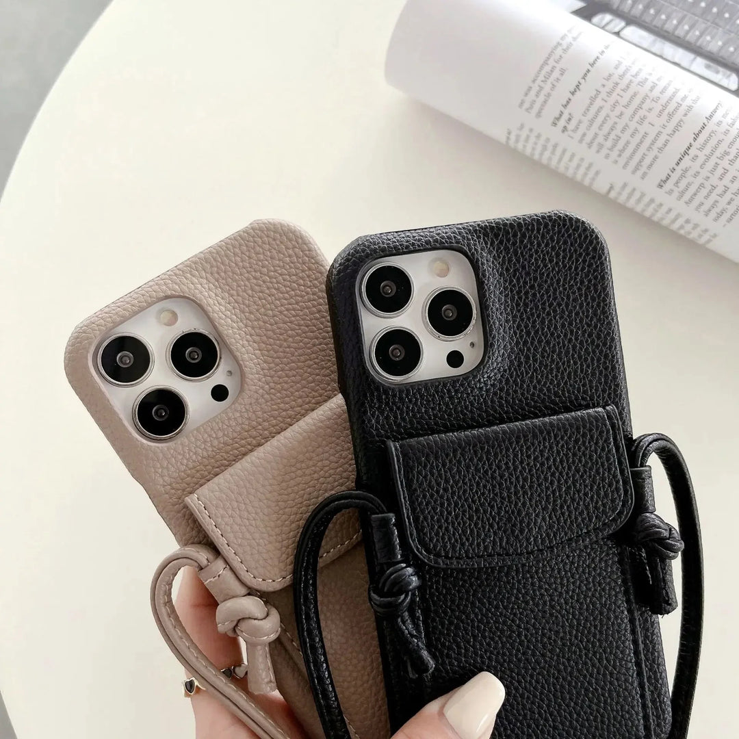 Two hands hold leather phone cases with detachable lanyards—one beige, one black—each featuring an attached mini pouch and wrist strap, over a white table with an open magazine in the background.