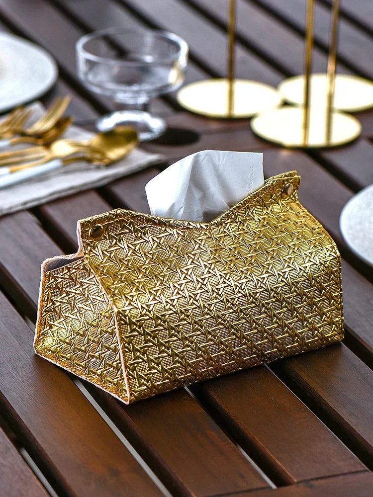 A gold, geometric tissue box adds a touch of luxury to the wooden table, surrounded by gold cutlery, plates, and glassware in an elegant dining setting.