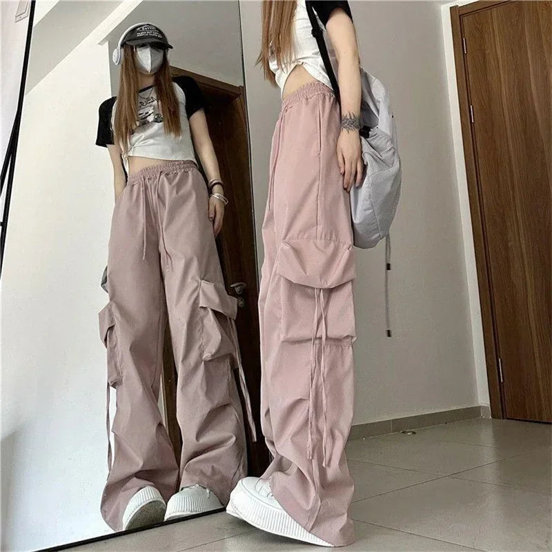 A person in high-waist pink cargo pants with cargo pockets, a black and white crop top, white sneakers, a baseball cap, a face mask, and a backpack stands before a mirror in a minimal room with tiled floor.