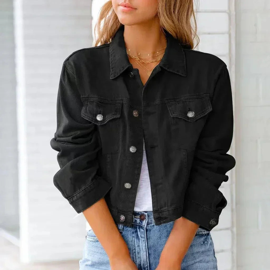 A woman wearing a black, button-up denim jacket over a white top and light blue jeans stands with her hands clasped in front of her, facing slightly to the side. She has long, wavy hair and is accessorized with layered necklaces.