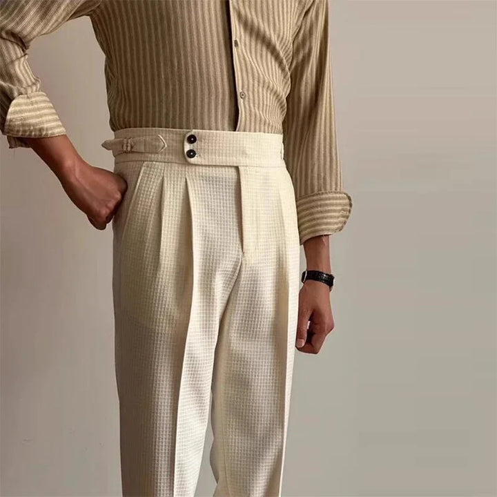 A person wearing a beige striped shirt and high-waisted, luxury tailored pants stands against a plain light-colored background. One hand is in their pocket, and they are accessorized with a dark wristwatch.