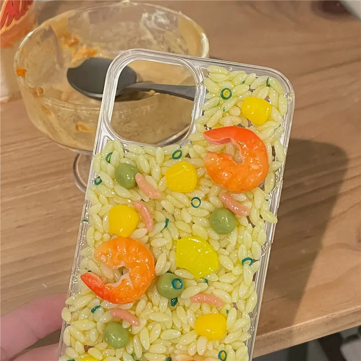 A clear phone case with realistic 3D detailing of rice, shrimp, peas, and corn offers rugged phone protection. It's held in front of a table with a used dessert bowl and spoon in the background.