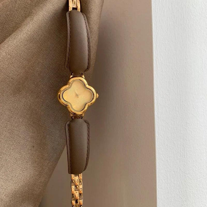 A luxury gold-plated clover dial watch with brown genuine leather strap is displayed against a beige fabric and white wall background.