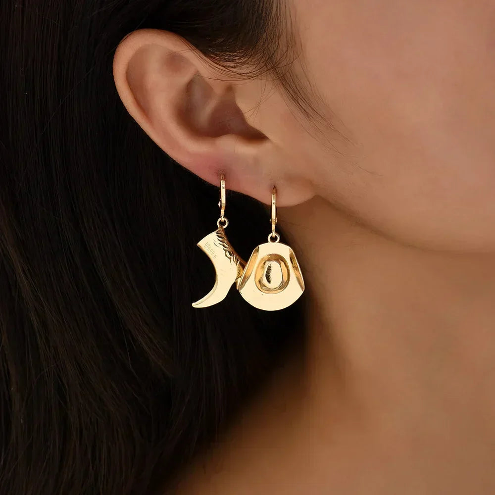 A close-up of a person’s ear wearing mismatched gold cowboy hat earrings and a boot, stylishly showcasing western jewelry against a neutral background.