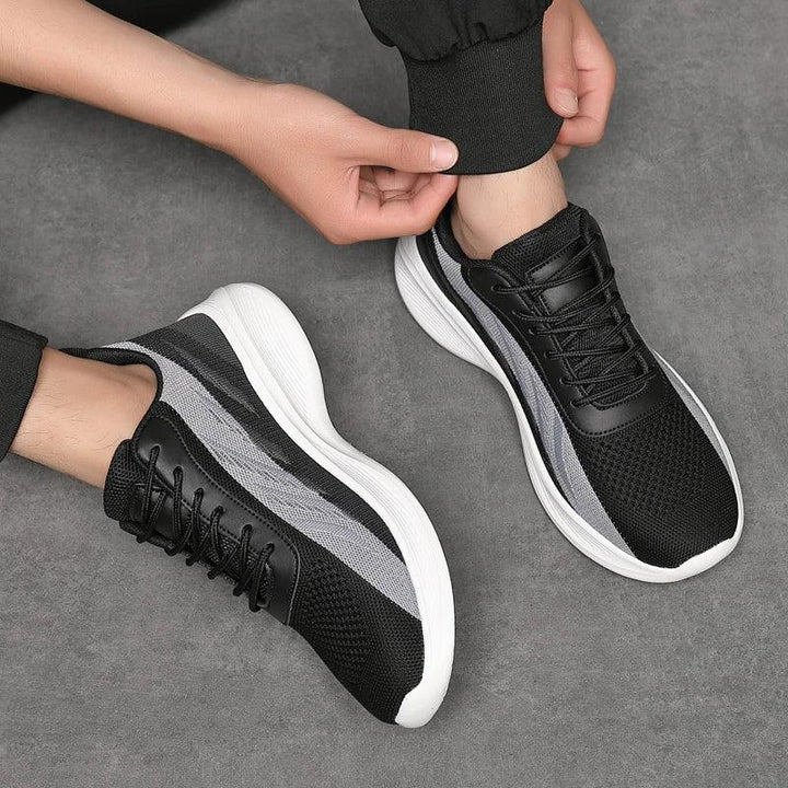 A person wearing black and gray athletic sneakers with breathable mesh and white soles sits on a gray surface, adjusting the cuff of their pants. Only the hands, feet, and lower legs are visible.