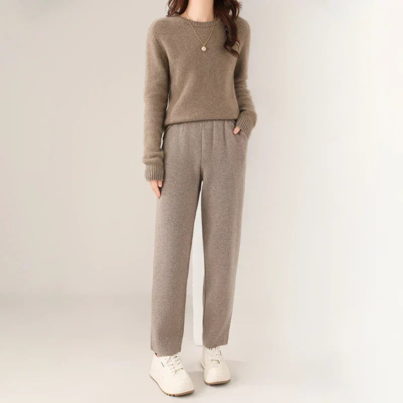 A woman wearing a brown sweater with ultra soft insulation, gray high-waisted pants featuring a slim tailored leg, and white sneakers stands against a plain light background. She has a necklace and her hands in her pockets. Her face is not visible.