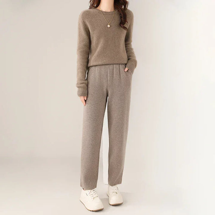 A woman wearing a brown sweater with ultra soft insulation, gray high-waisted pants featuring a slim tailored leg, and white sneakers stands against a plain light background. She has a necklace and her hands in her pockets. Her face is not visible.